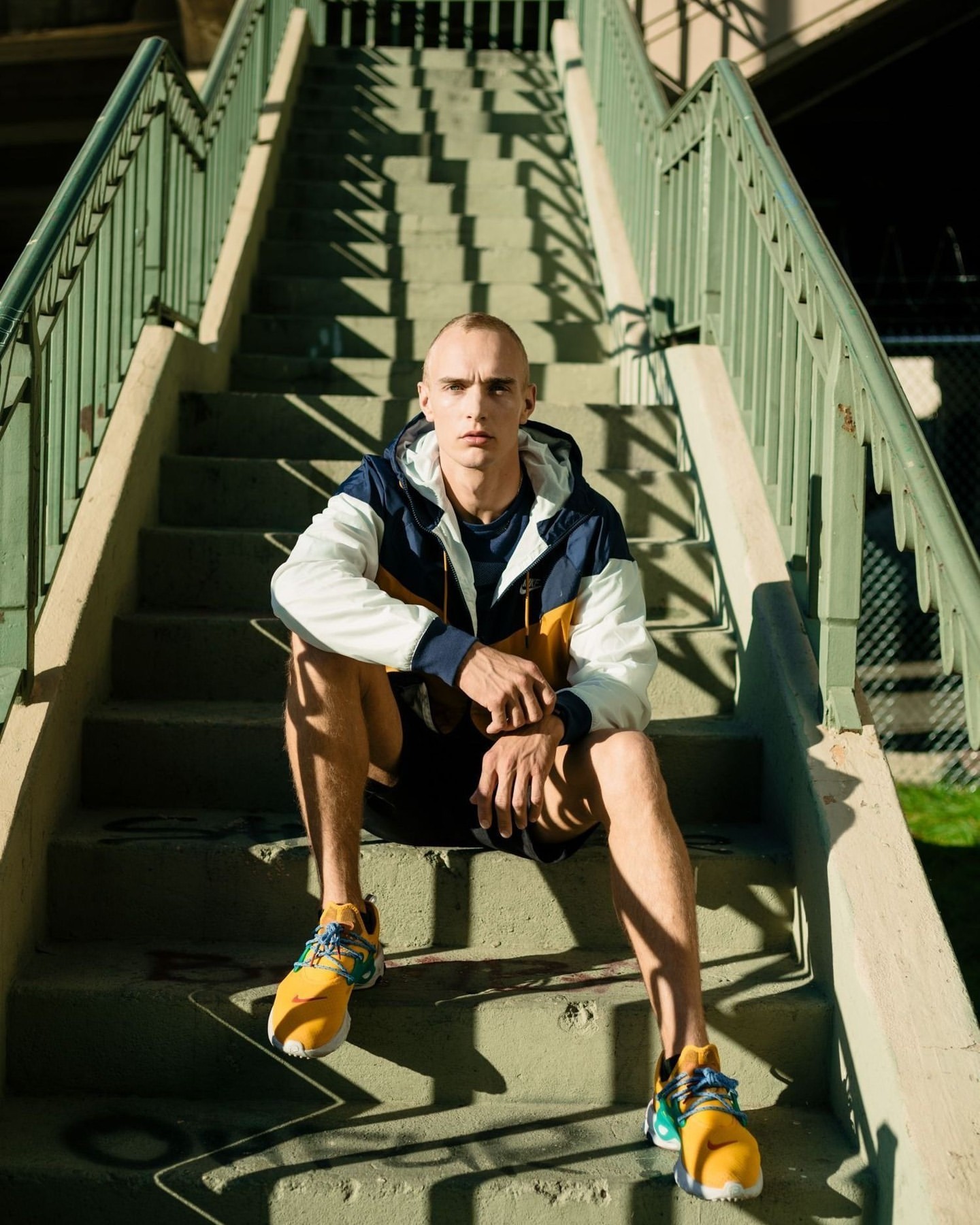 Adult male in a windbreaker and athletic shoes sitting on an outdoor staircase with green railing, creating linear shadows.