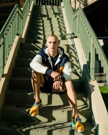 Adult male in a windbreaker and athletic shoes sitting on an outdoor staircase with green railing, creating linear shadows.