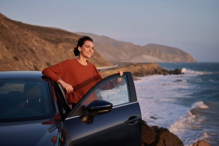 Woman leans against a car door with the ocean, mountains, and blue sky in the background, captured during daylight at the beach.