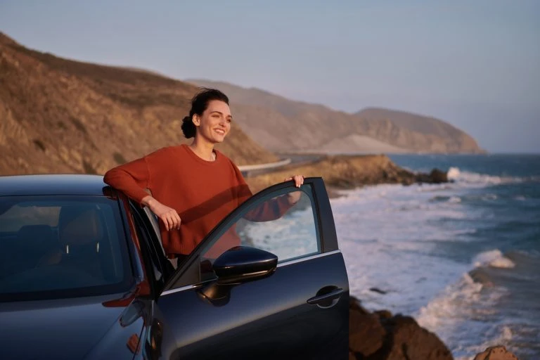 Woman leans against a car door with the ocean, mountains, and blue sky in the background, captured during daylight at the beach.