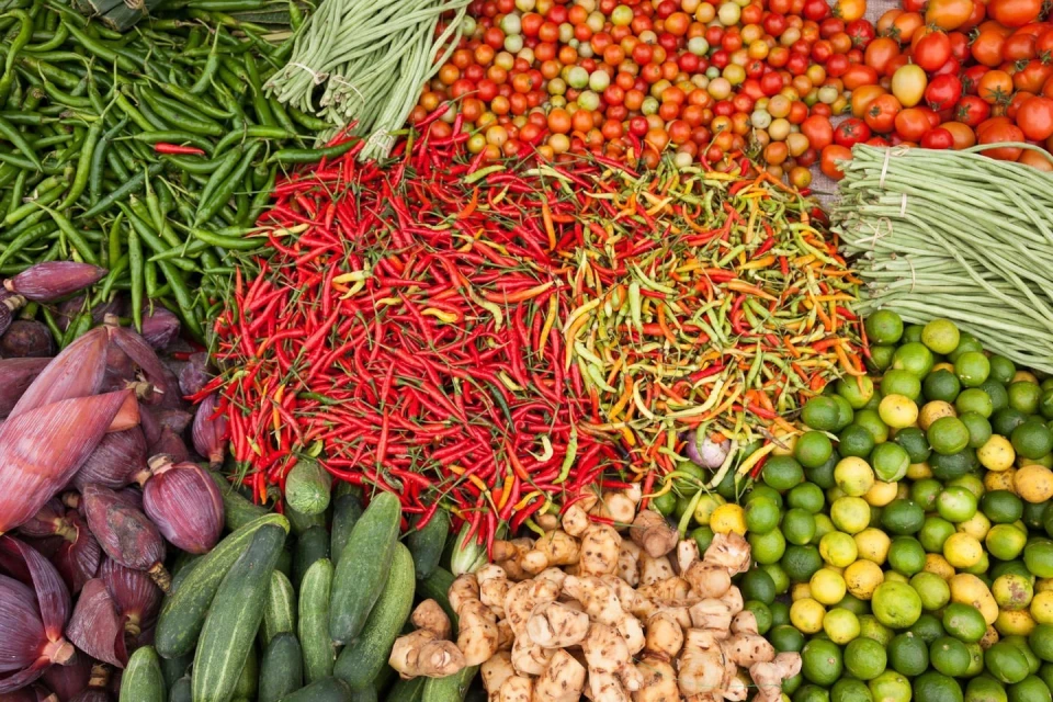 Overhead shot displays colorful assortment of red & green chilies, tomatoes, limes, cucumbers, ginger, & banana flowers for food photography.