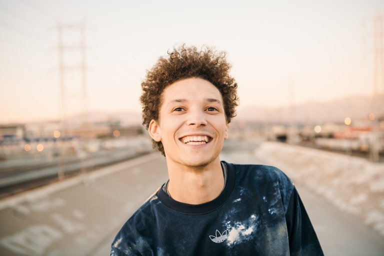 Smiling adult male with curly hair wearing an Adidas sweatshirt outside on an overcast day. Cityscape and powerlines in the background.