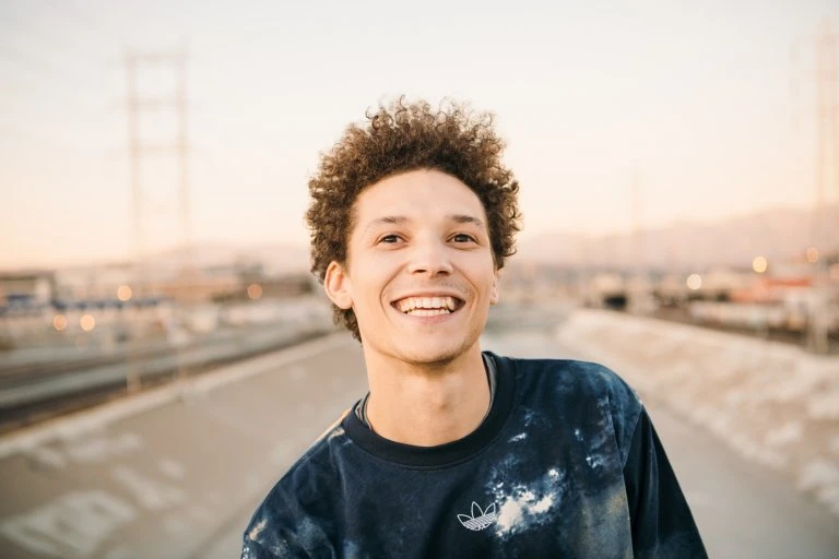 Smiling adult male with curly hair wearing an Adidas sweatshirt outside on an overcast day. Cityscape and powerlines in the background.