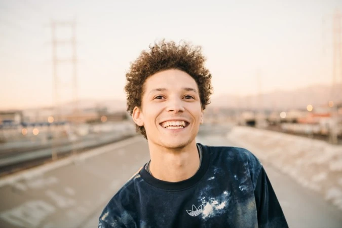 Smiling adult male with curly hair wearing an Adidas sweatshirt outside on an overcast day. Cityscape and powerlines in the background.