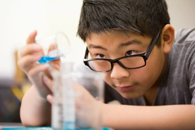 Young boy wearing black glasses pouring blue liquid from a small glass beaker into a graduated cylinder during a science activity