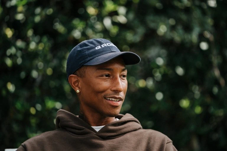 Close-up of an adult male wearing a baseball cap and a brown hoodie, smiling slightly against a blurred green foliage background.