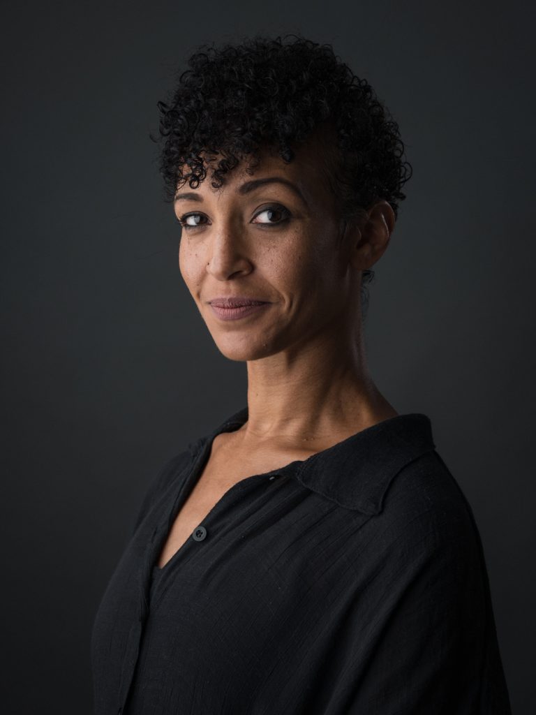 Portrait of a smiling adult female with curly hair and a black button-down shirt, set against a dark gray background in a photo studio.