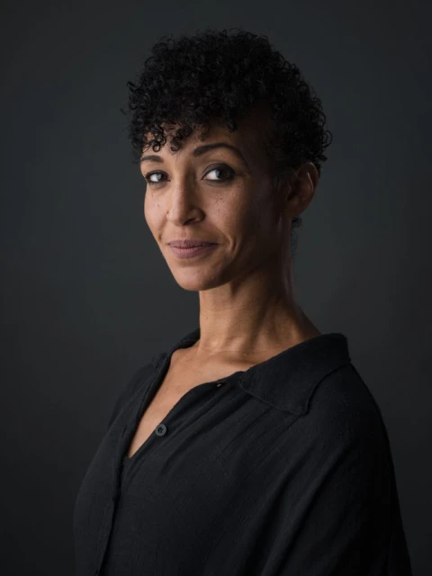 Portrait of a smiling adult female with curly hair and a black button-down shirt, set against a dark gray background in a photo studio.