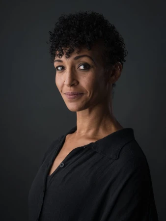 Portrait of a smiling adult female with curly hair and a black button-down shirt, set against a dark gray background in a photo studio.