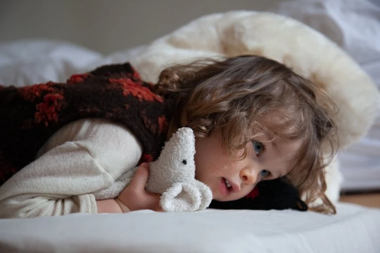 Child with curly hair rests on a white bed, holding a stuffed animal, wearing a brown and red vest and a cream long-sleeve shirt.