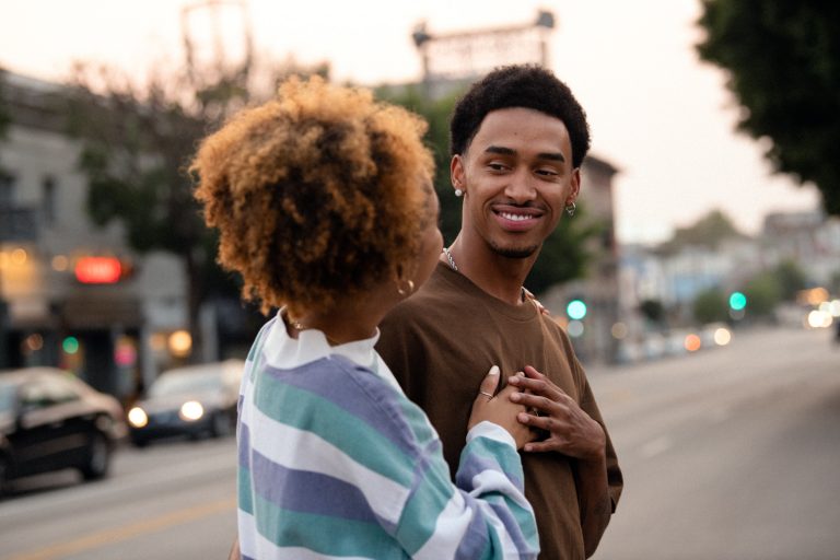 A young couple holding hands on a city street at dusk, with buildings and cars in the background, and soft, warm lighting.