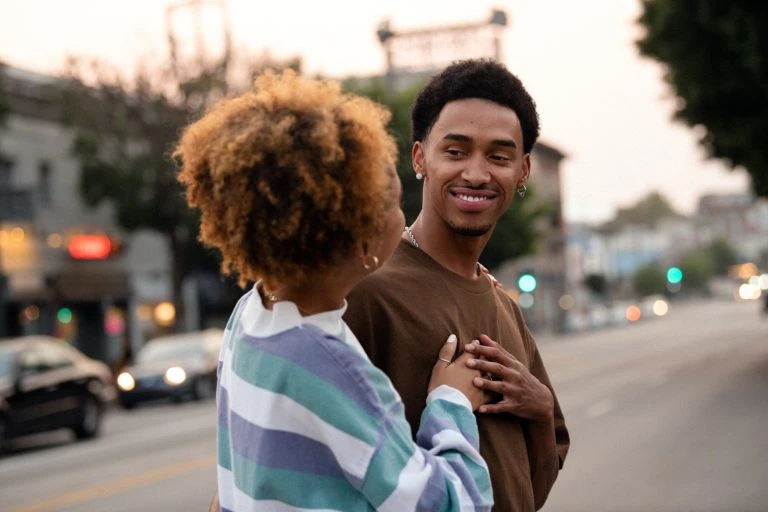 A young couple holding hands on a city street at dusk, with buildings and cars in the background, and soft, warm lighting.