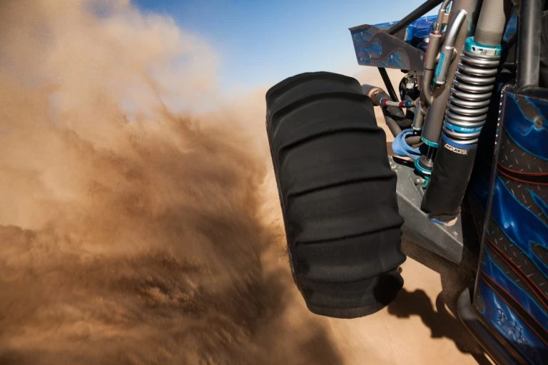 Close-up of a custom off-road vehicle's tire kicking up dust in an outdoor desert environment against a blue sky.