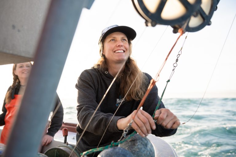 Woman working on a fishing boat on the ocean, wearing a sweatshirt and baseball cap while pulling ropes connected to overhead rigging.