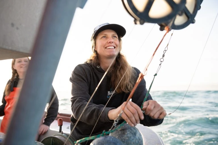 Woman working on a fishing boat on the ocean, wearing a sweatshirt and baseball cap while pulling ropes connected to overhead rigging.