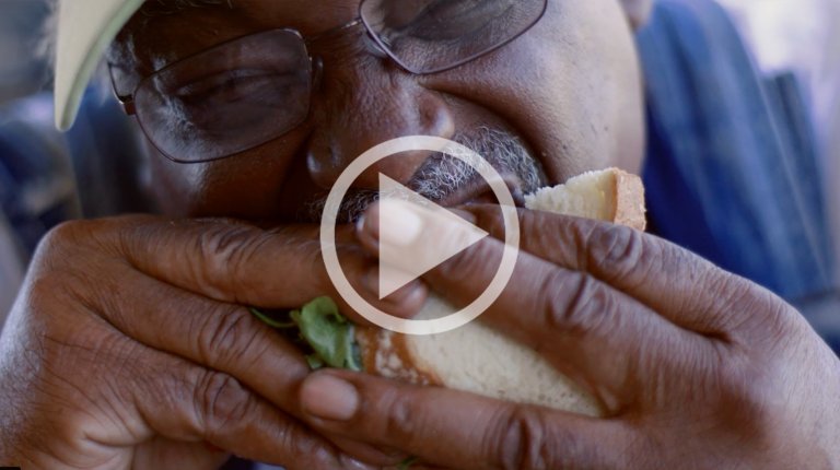 Close-up of a man wearing glasses eating a large sandwich with white bread and green filling, photographed by Peter Bohler.