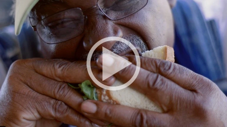 Close-up of a man wearing glasses eating a large sandwich with white bread and green filling, photographed by Peter Bohler.