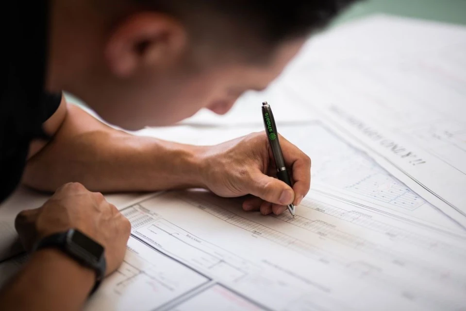Person writing on paper with a pen, wearing a black smartwatch. The paper contains technical diagrams and text.