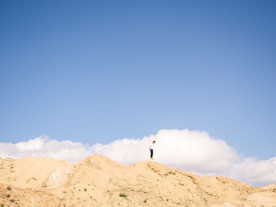 A lone person stands on a sandy hill under a vast blue sky with white clouds, photographed by Peter Bohler.