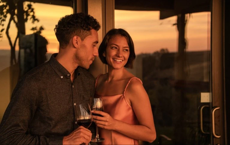 Smiling couple toasting with red wine indoors at sunset. They stand by a glass door with a view of trees in the background.