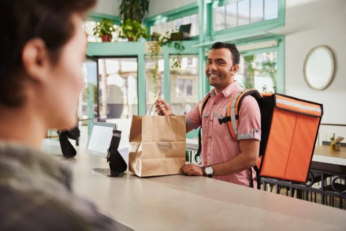 Delivery courier with orange backpack handing paper bag to customer at counter