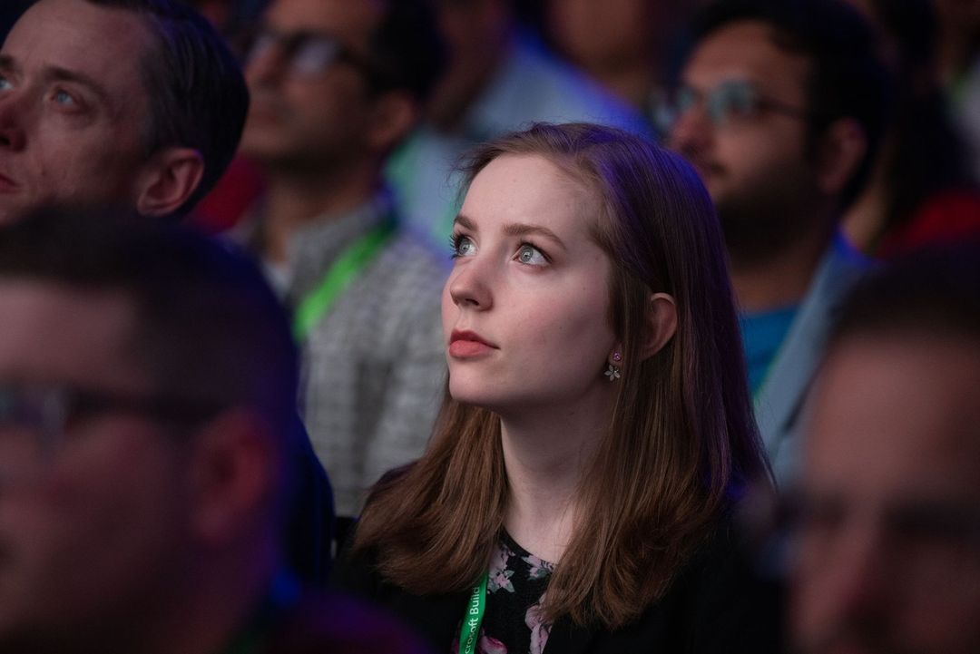 Attentive woman in a crowd, looking up with a slight smile, surrounded by blurred faces in an indoor setting with soft, even lighting.