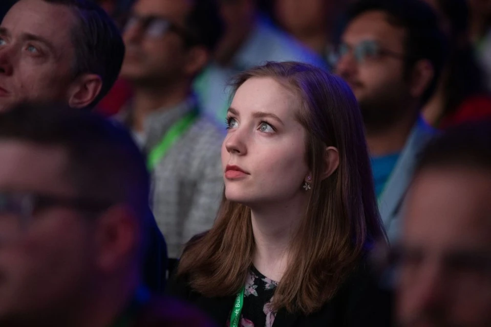Attentive woman in a crowd, looking up with a slight smile, surrounded by blurred faces in an indoor setting with soft, even lighting.