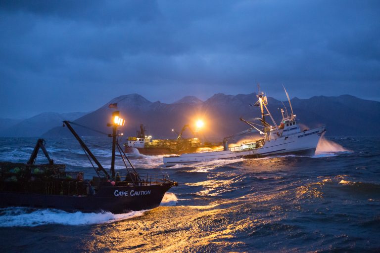 Fishing boats navigate choppy ocean water near mountainous landmass under blue, cloudy skies during dusk or dawn with boat lights on.