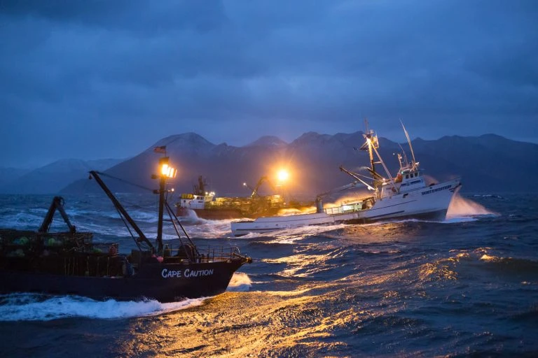 Fishing boats navigate choppy ocean water near mountainous landmass under blue, cloudy skies during dusk or dawn with boat lights on.