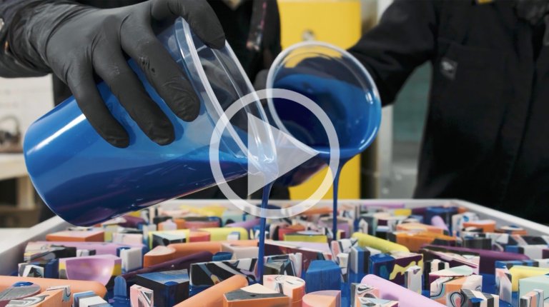 Two gloved hands pour blue liquid from clear containers onto an arrangement of colorful geometric shapes inside a white tray.