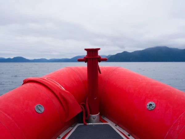 Inflatable boat on water with distant mountains beneath an overcast sky. The red inflatable boat dominates the foreground view.