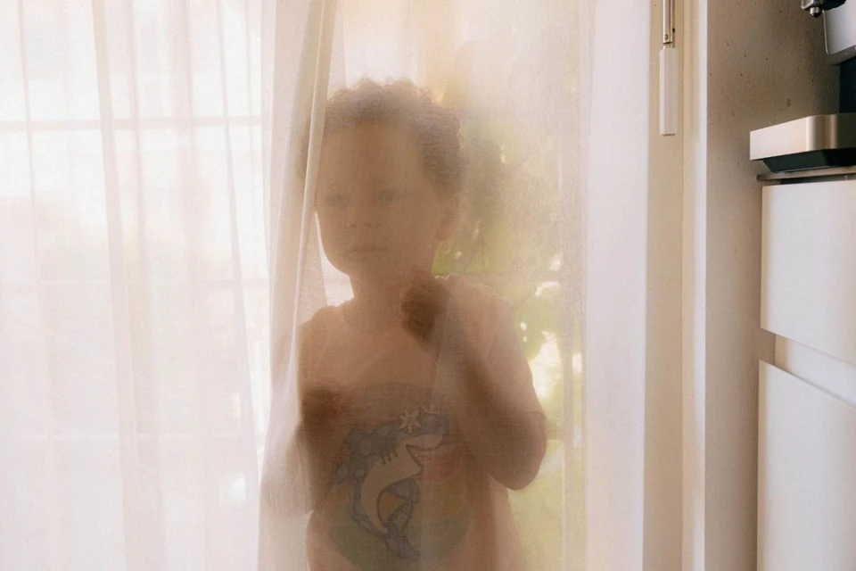 Young child stands behind sheer white curtains in an indoor setting, creating a soft, diffused view of the child's form and features.