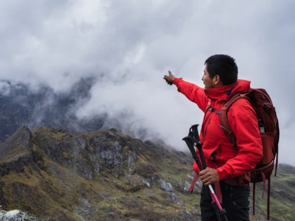 Adventurer in a red jacket points over a mountain ridge shrouded in clouds, holding trekking poles with a backpack, nature scene.