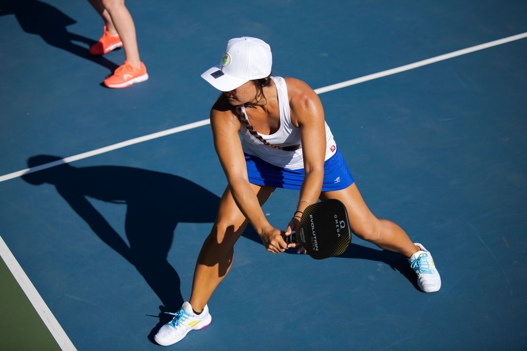 A woman plays pickleball on an outdoor court, wearing a white cap, tank top, and blue skirt, holding a paddle, viewed from overhead.