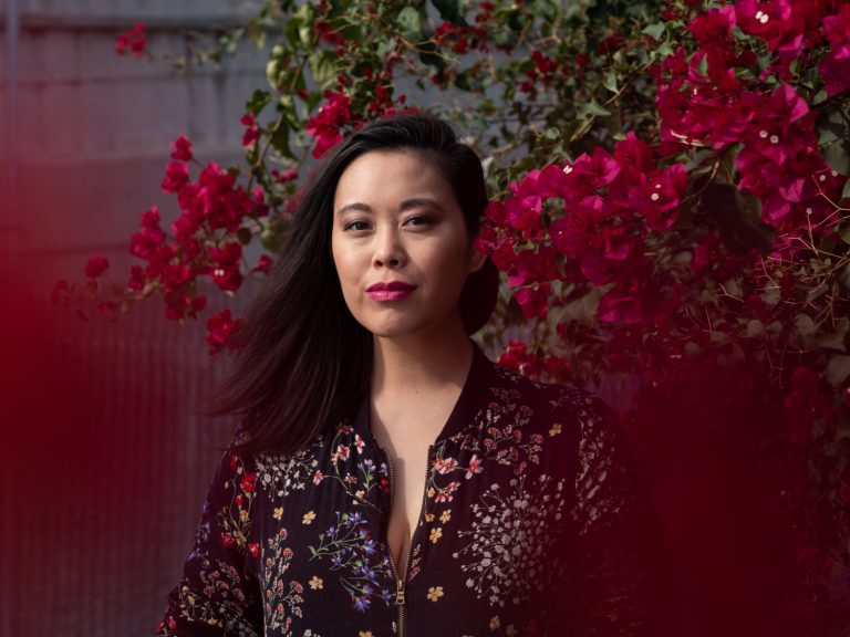 Portrait of an Asian woman with a floral jacket. She stands outdoors with red flowers and blurred red foreground.