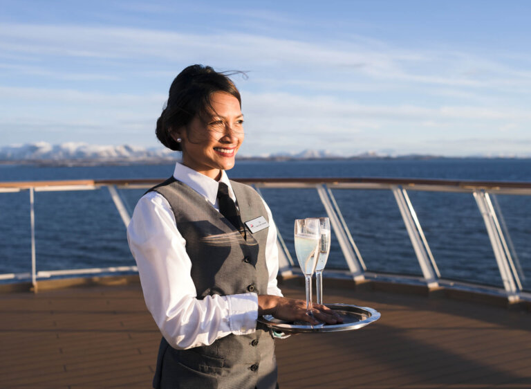 Smiling server woman in gray uniform holding champagne glasses on cruise ship with the ocean and distant mountains on a sunny day.