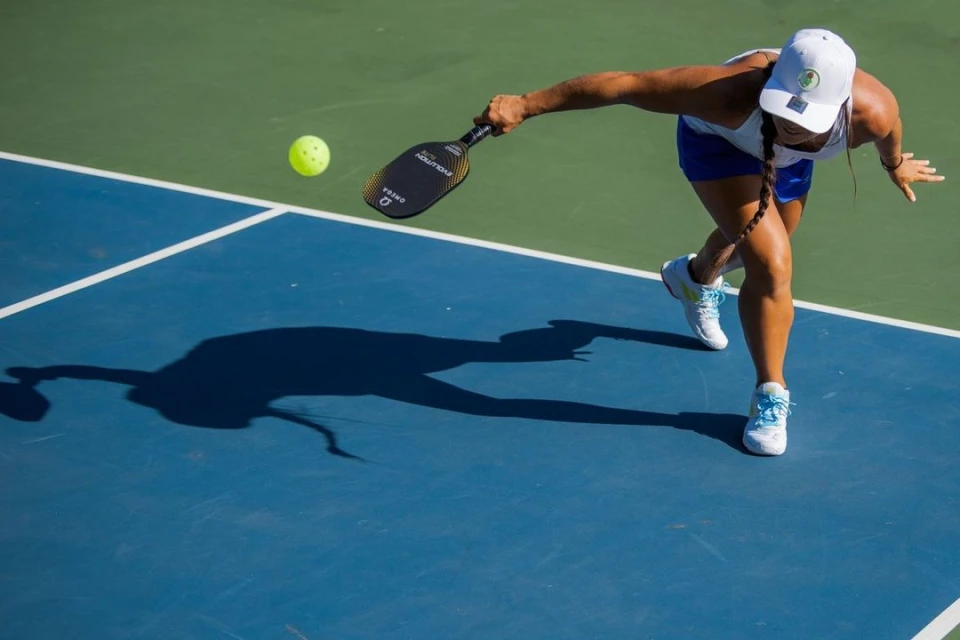 Action shot of an adult female pickleball player in white hat swinging a paddle at a yellow ball on a blue court, outdoors.