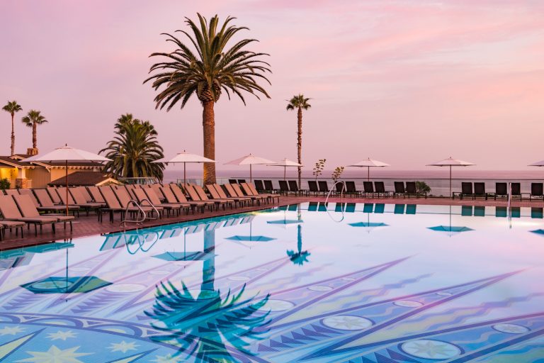 View of an outdoor swimming pool with lounge chairs and palm trees at sunset. The pool reflects the palm trees and pink-hued sky.