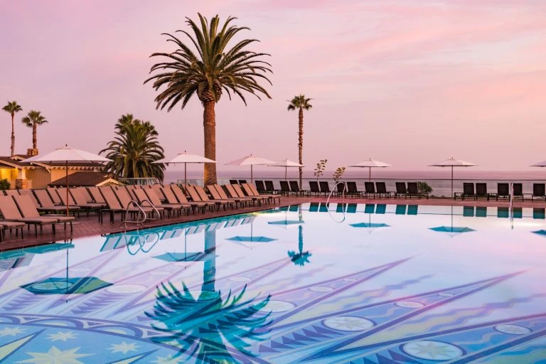 View of an outdoor swimming pool with lounge chairs and palm trees at sunset. The pool reflects the palm trees and pink-hued sky.