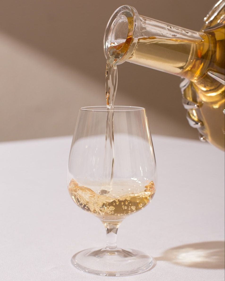 Golden amber liquid being poured from a decanter into a clear stemmed glass on a white tablecloth