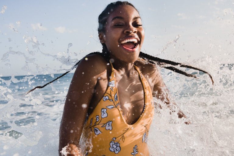 Smiling African-American woman in floral swimsuit enjoys the ocean's splash against a blue sky, capturing a vibrant summer beach moment.