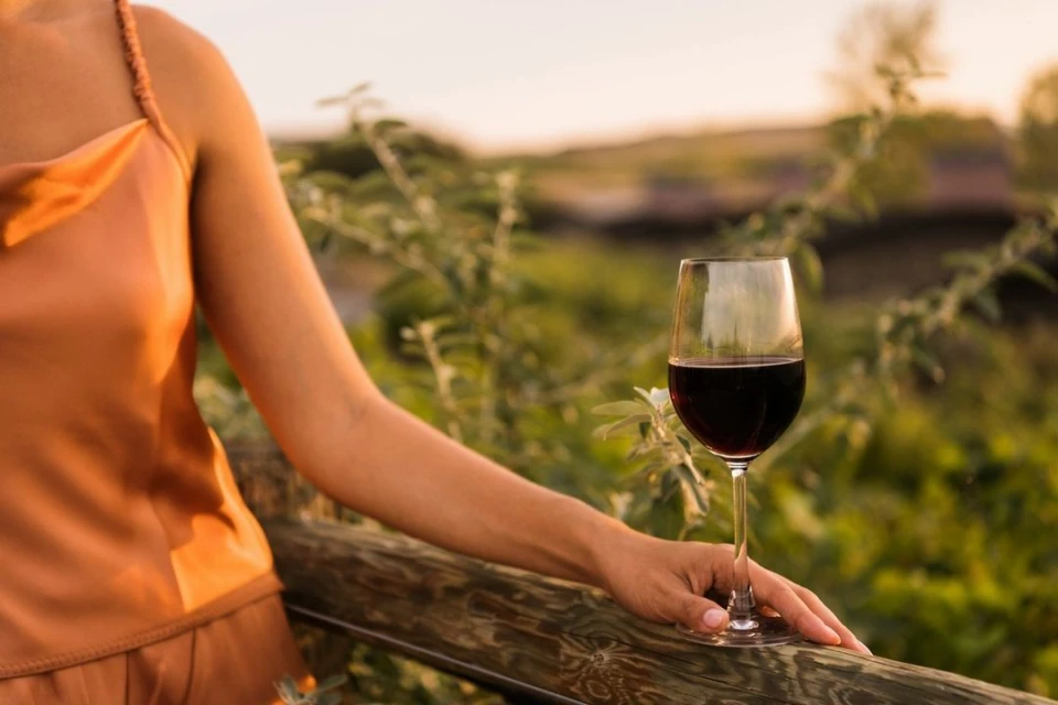 Woman in peach-colored top holds a wine glass filled with red wine, resting her hand on a wooden rail in an outdoor setting.