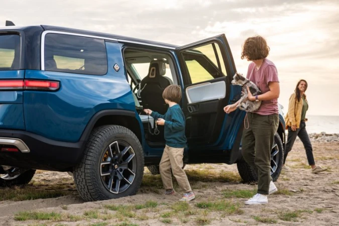 Family of three and a dog near a blue Rivian electric SUV on a sandy beach with the ocean and sky visible in the background.