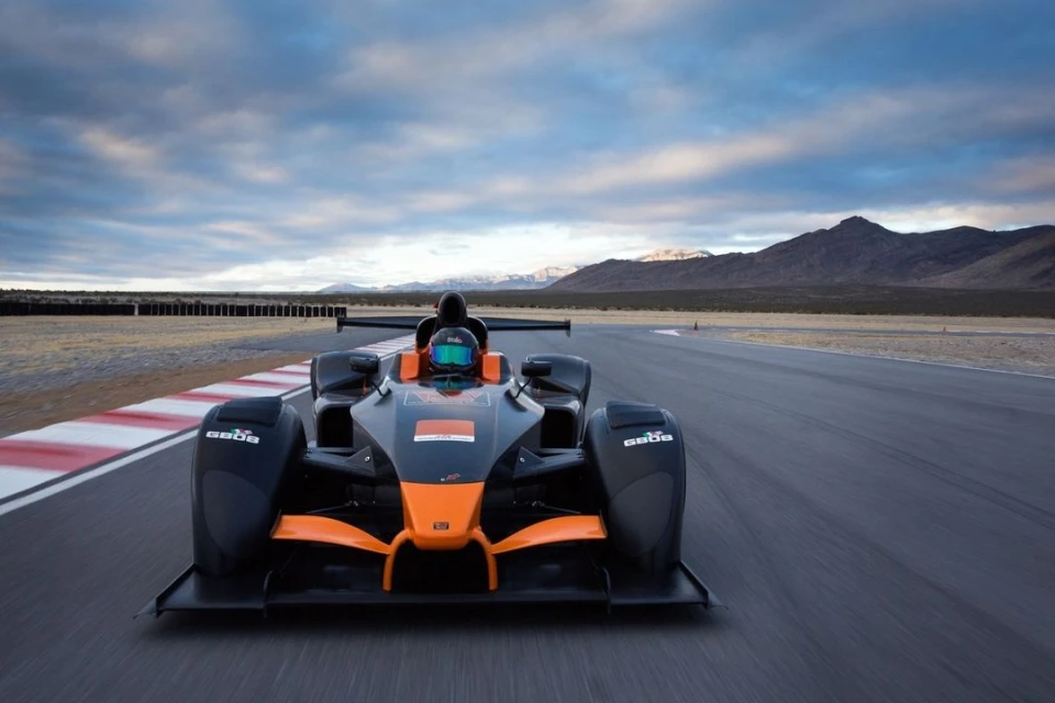 Front view of a black and orange open-wheel race car driving on a track on a sunny day with mountains and a blue sky in the background.