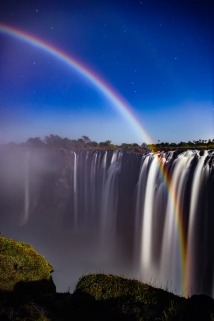 Majestic Victoria Falls captured at night, a rainbow arches across the sky under a starlit blue sky, with mist rising from the falls.