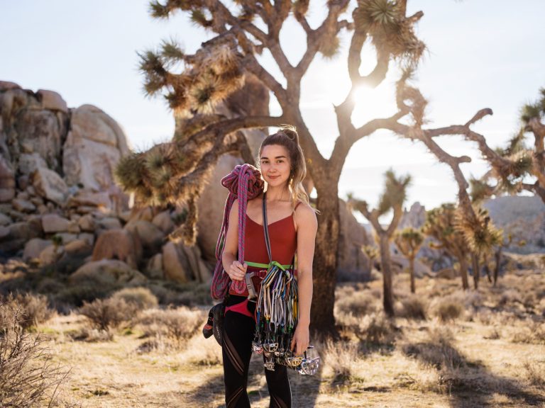 A woman with climbing gear stands in a desert landscape with Joshua trees, wearing a maroon top and black leggings, smiling at the camera.
