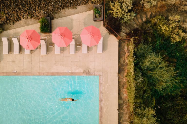 Aerial view of woman swimming in outdoor pool, surrounded by lounge chairs under pink umbrellas, near lush greenery and stone steps.