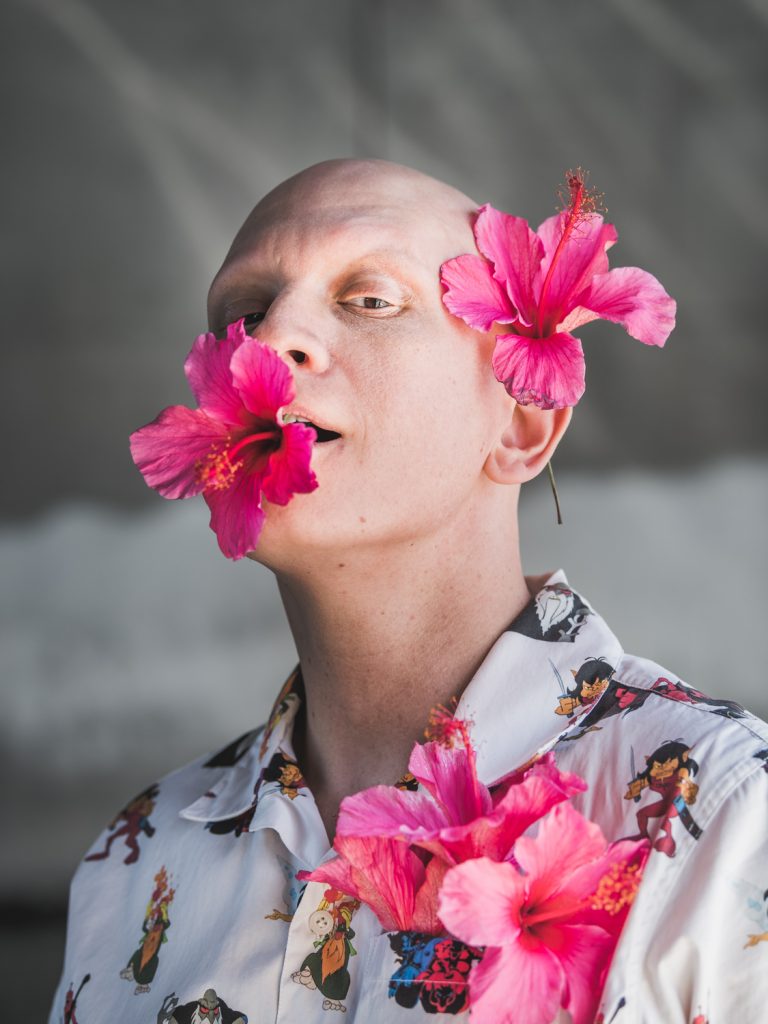 Close-up of a bald person with pink hibiscus flowers in their mouth, behind their ear, and on their patterned shirt.