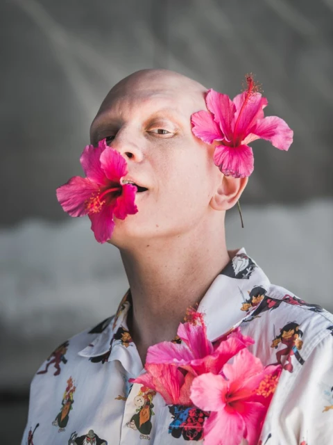 Close-up of a bald person with pink hibiscus flowers in their mouth, behind their ear, and on their patterned shirt.