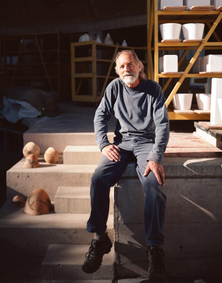 Mature man with a beard sits on concrete steps outdoors, wearing a sweatshirt and jeans, with pottery shelves visible in the background.